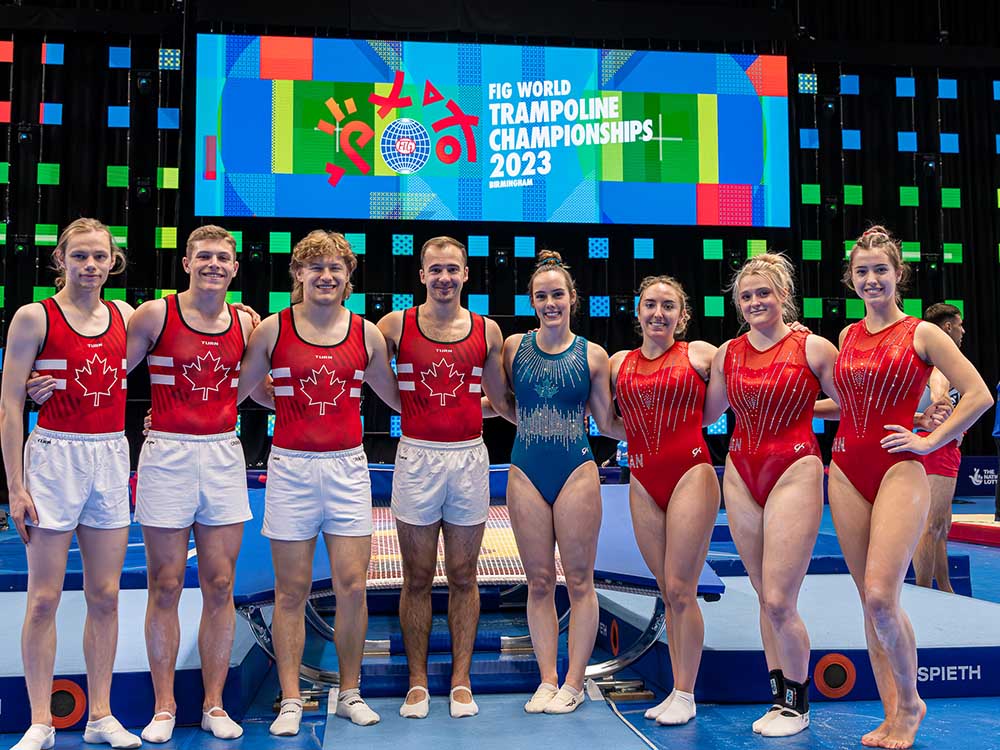 A group of 8 gymnasts stands in front of a sign that says "FIG World Trampoline Championships 2023"